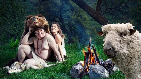 caveman-type people with animal skins, sitting next to a fire with a sheep